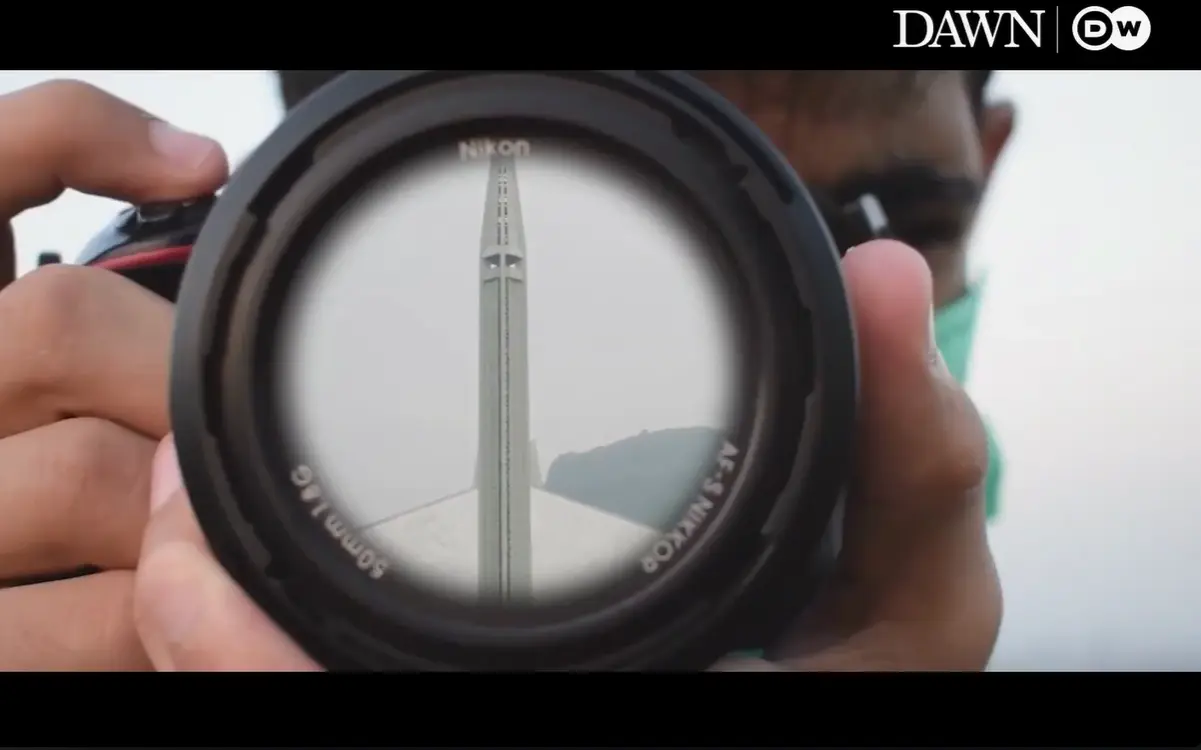 A still from the film: someone holds a camera lens up to their eye, and a tall minaret stands framed inside it against a pale sky. The Dawn and DW logos sit in the corner.